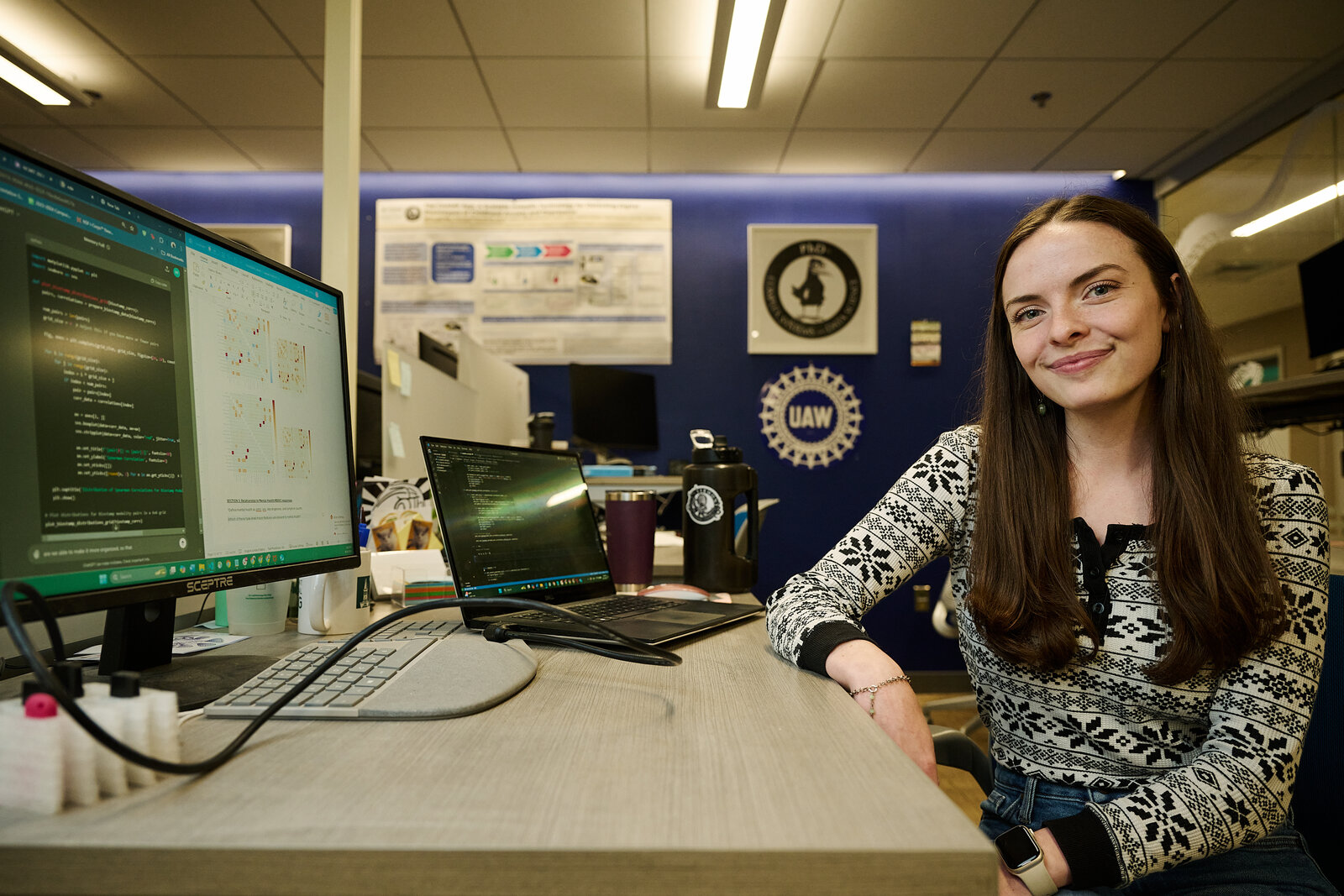 Bryn at her PhD workstation with code, figures, and a research poster on the wall.